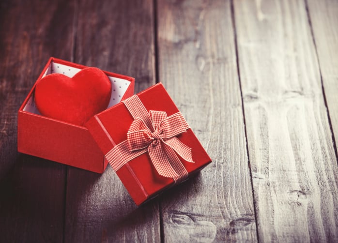 Red gift box with toy heart inside on wooden table.