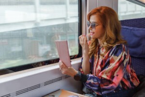 Beautiful fashionable mid adult woman sitting in window seat in day train, going downtown and putting lip gloss on, using smart phone as a mirror: Travel Beauty Essentials