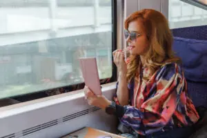 Beautiful fashionable mid adult woman sitting in window seat in day train, going downtown and putting lip gloss on, using smart phone as a mirror: Travel Beauty Essentials