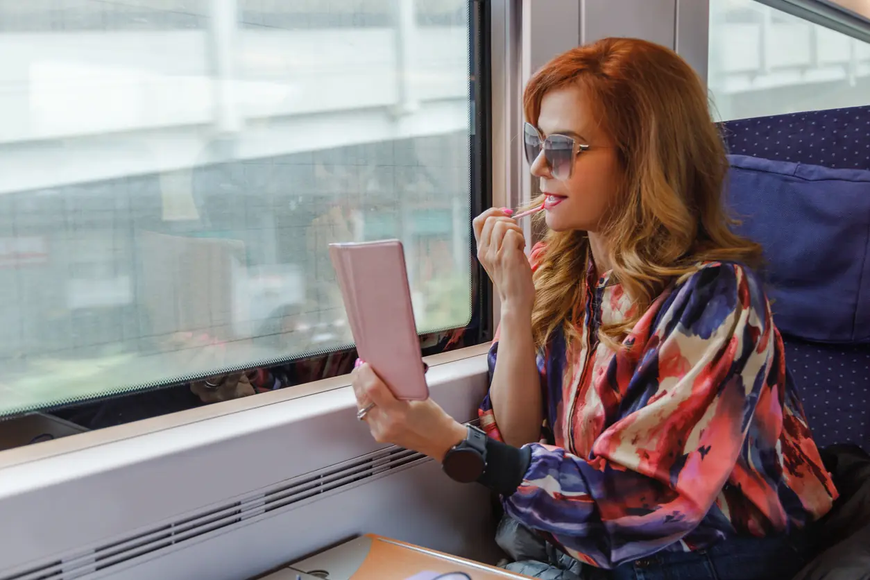 Beautiful fashionable mid adult woman sitting in window seat in day train, going downtown and putting lip gloss on, using smart phone as a mirror: Travel Beauty Essentials