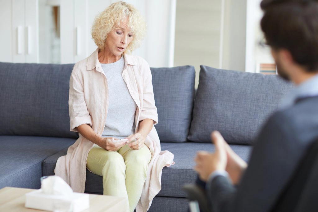  mature woman with curly hair holding napkin and nervously sharing her problems without looking psychiatrist in eye at psychotherapy session for emotional health
