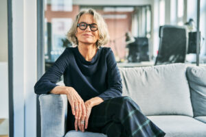 Best Colors to Wear for Gray Hair -- Portrait of a confident senior businesswoman sitting on a sofa in an office lobby, looking at the camera with a warm and professional smile