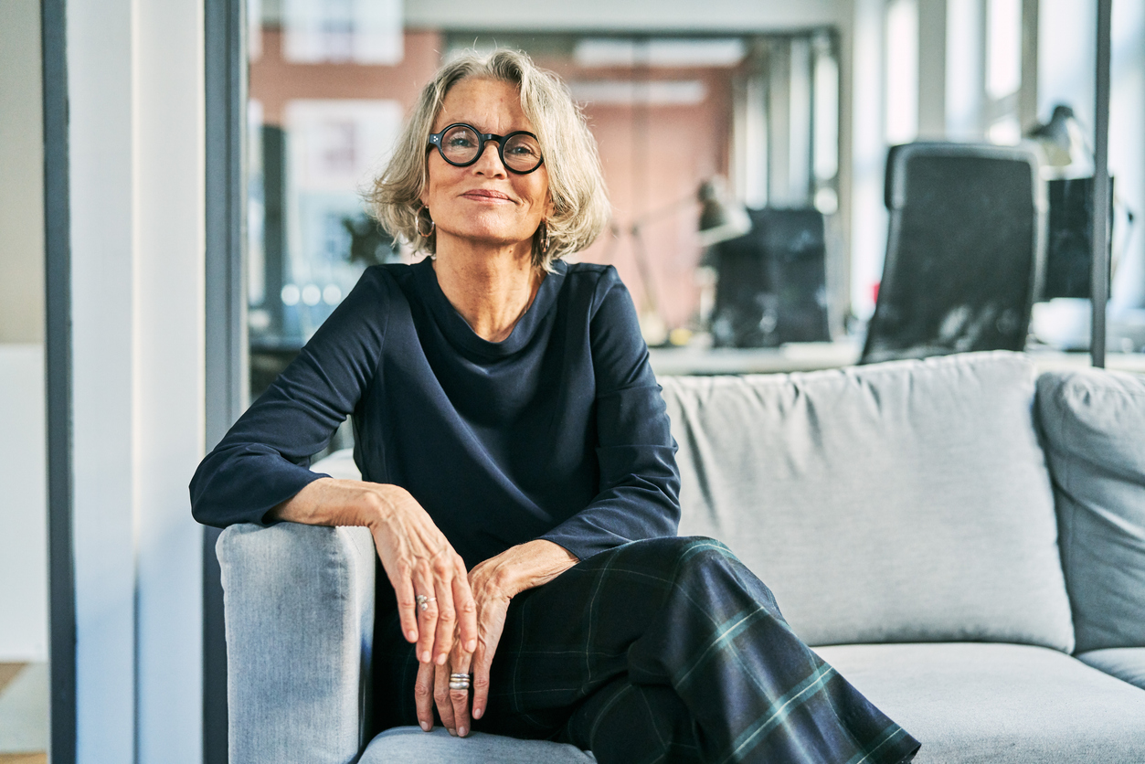 Best Colors to Wear for Gray Hair -- Portrait of a confident senior businesswoman sitting on a sofa in an office lobby, looking at the camera with a warm and professional smile