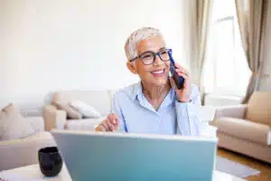 Smiling mature beautiful business woman with white hair working on laptop in bright modern home office. Business woman talking on her mobile phone while working from home