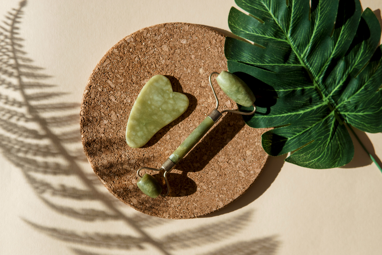 10 Best Skincare Tools for Mature Skin -- Jade Gua sha scraper and face roller massager on a cork round stand with a monstera leaf. Hard light, shadows, the concept self-care. Facial care. Zero waste. Lifting and toning treatment at home.