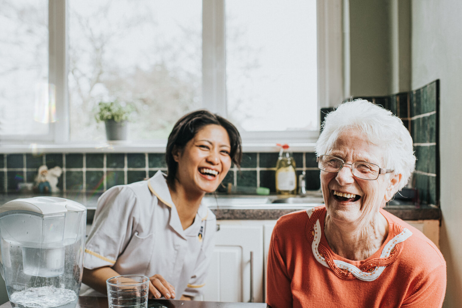 An elderly woman laughs beside a friendly young care assistant
