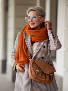 Smiling Woman Outdoor Portrait. Short blonde hair fashion model wears stylish clothes, double-breasted jacket, leather handbag, ochre knitted scarf and glasses. Fashion trend of autumn or spring