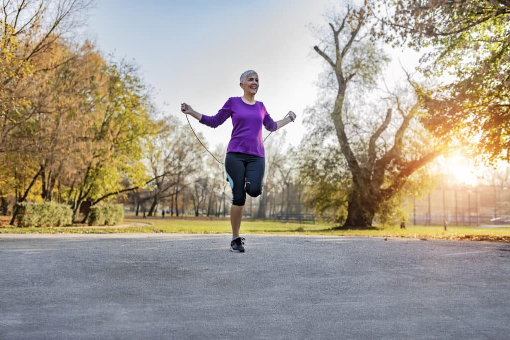 Happy Senior Woman Exercising With Jump-rope Outdoors - Fitness, Sport, Training, Park and Lifestyle Concept. Elderly Fitness Woman Skipping With a Jump Rope Outdoors. Mature Female Doing Fitness Training in Morning in the Public Park