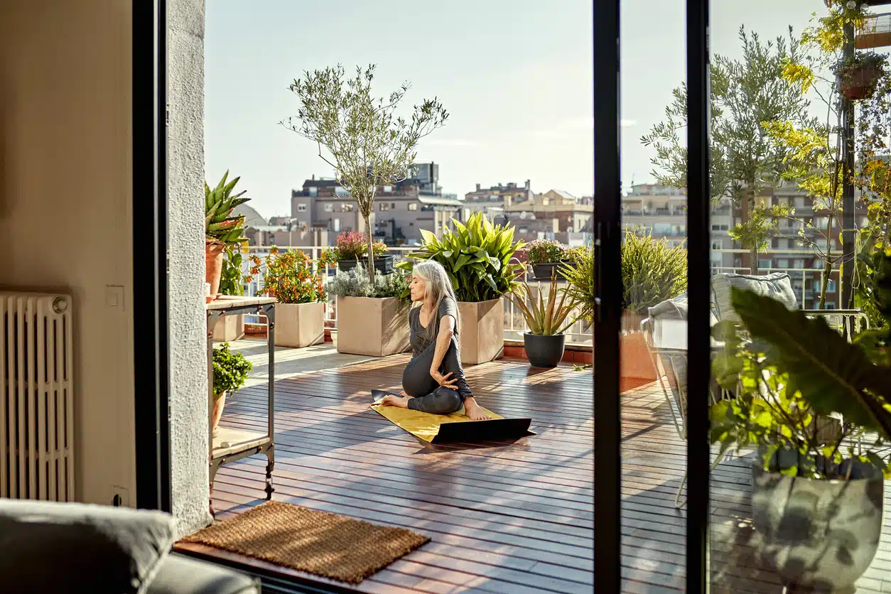 Full length front view of Caucasian woman with long gray hair photographed through open sliding glass door on exercise mat in seated spinal twist position.