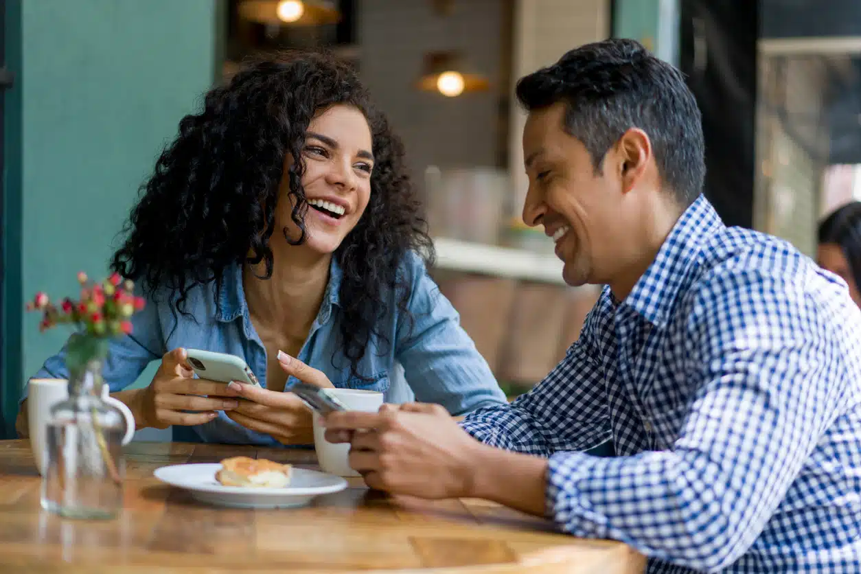 Happy Latin American couple on a date at a cafe looking at social media on their cell phones and laughing â lifestyle concepts