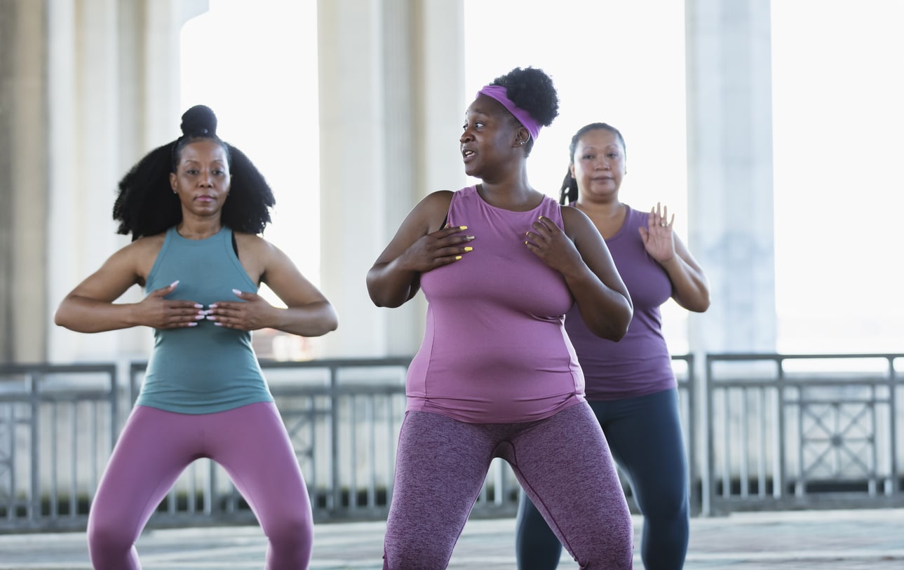 A multiracial group of three women exercising together outdoors under a bridge on a city waterfront. The two African-American women are in their 40s. Their Pacific Islander friend is in her 30s.