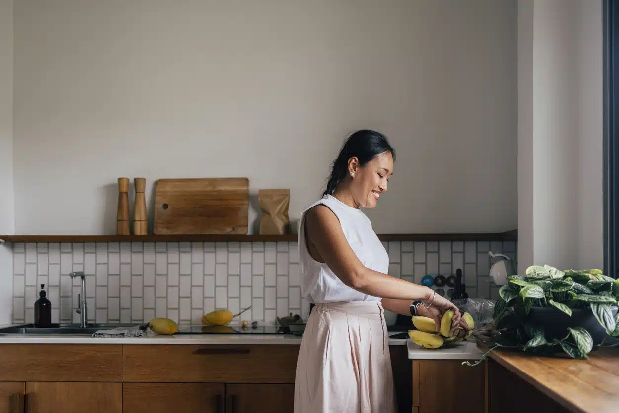 Horizontal photo of a young Asian woman taking a banana from a kitchen counter.