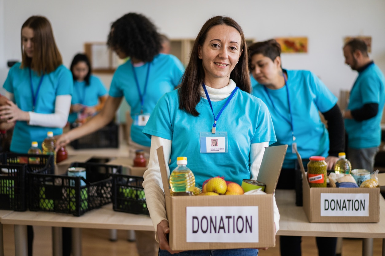 A female charity assistant is carrying a heavy box of donated groceries ready to be delivered to victims of poverty and hunger, in the background more volunteers are busy packing more boxes in a local food bank