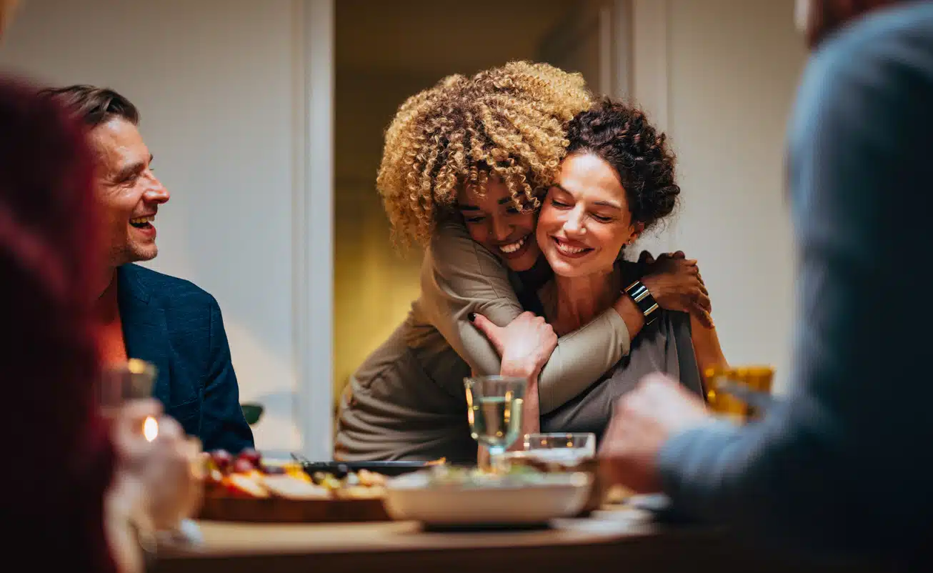 Cheerful smiling African-American woman embracing her female friend during a dinner with family and friends.