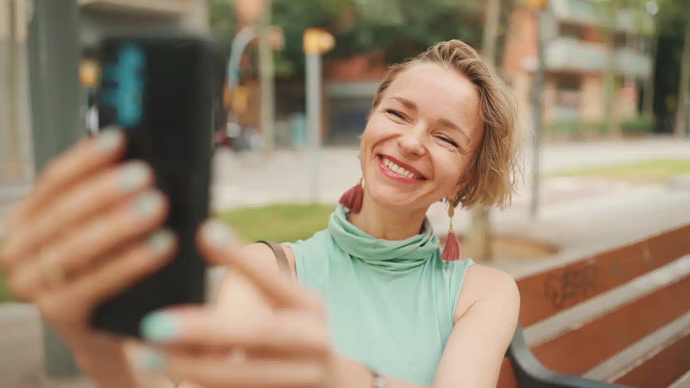 Beautiful happy smiling woman with short blond hair in casual wear sits on bench, makes video call on cellphone