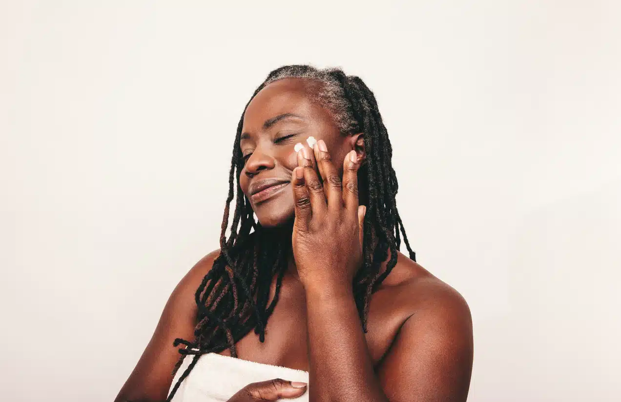 Mature woman with dreadlocks applying moisturizing cream on her face. Confident dark-skinned woman taking care of her flawless melanated skin. Mature black woman ageing gracefully.