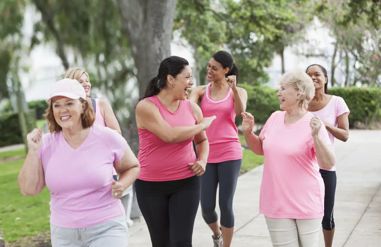 Multi-ethnic group of women (30s, 40s, 60s) wearing pink, participating in breast cancer walk. Focus on excited Hispanic woman (30s) in middle.