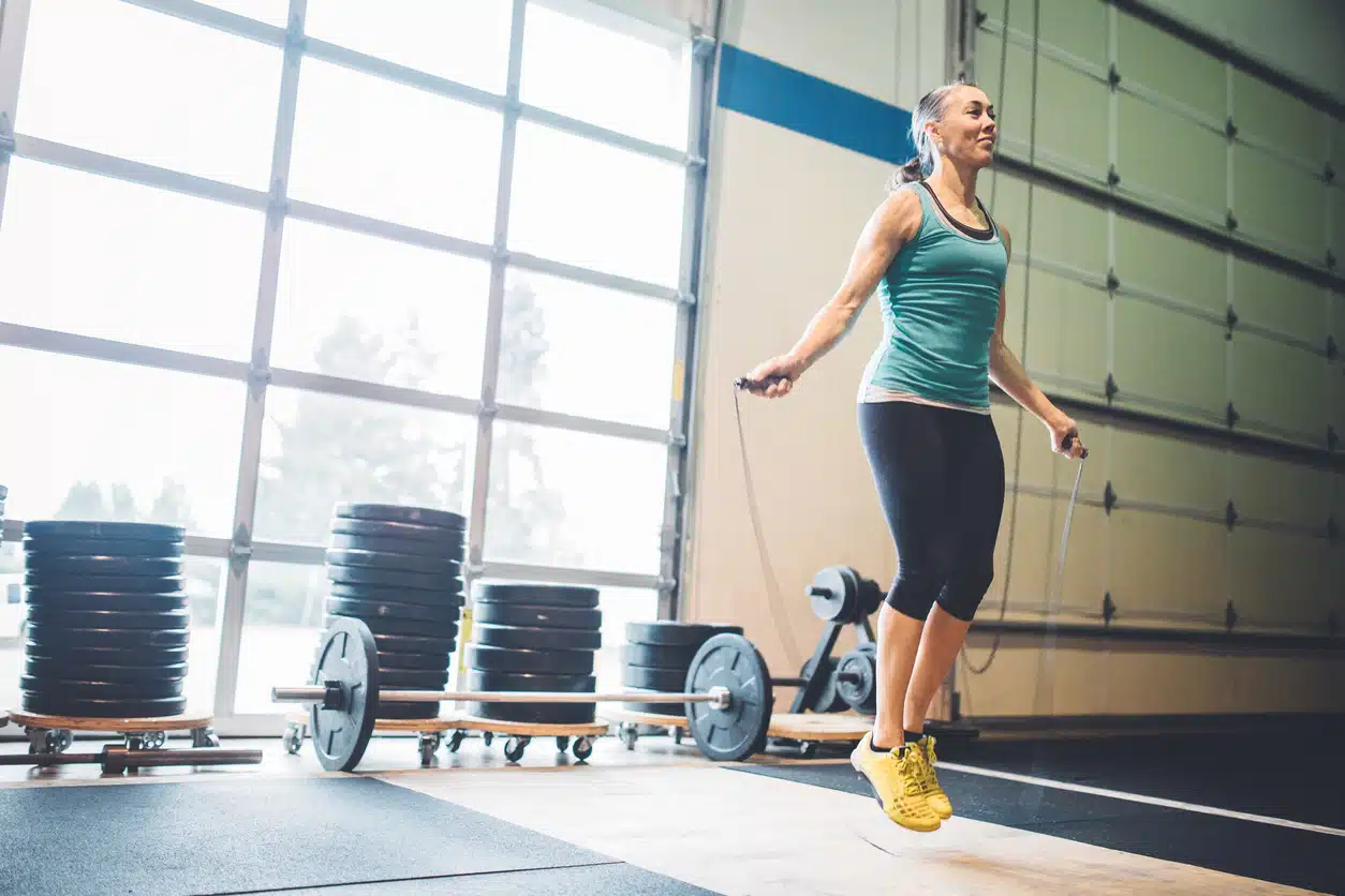 A healthy and fit woman in her 50's jumps rope with a smile on her face. Bright clean light shines in the window behind her in a cross training setting. 