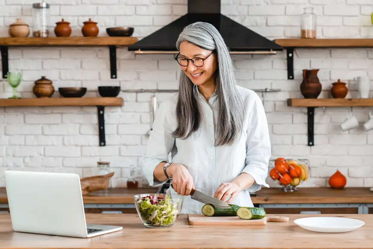Cheerful middle-aged woman cooking in the kitchen using laptop