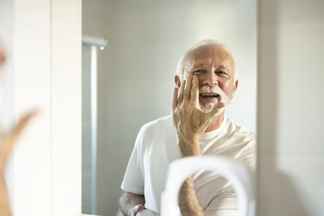 Skin care. Handsome bearded senior man applying cream at his face and looking at himself with smile while standing in front of the mirror