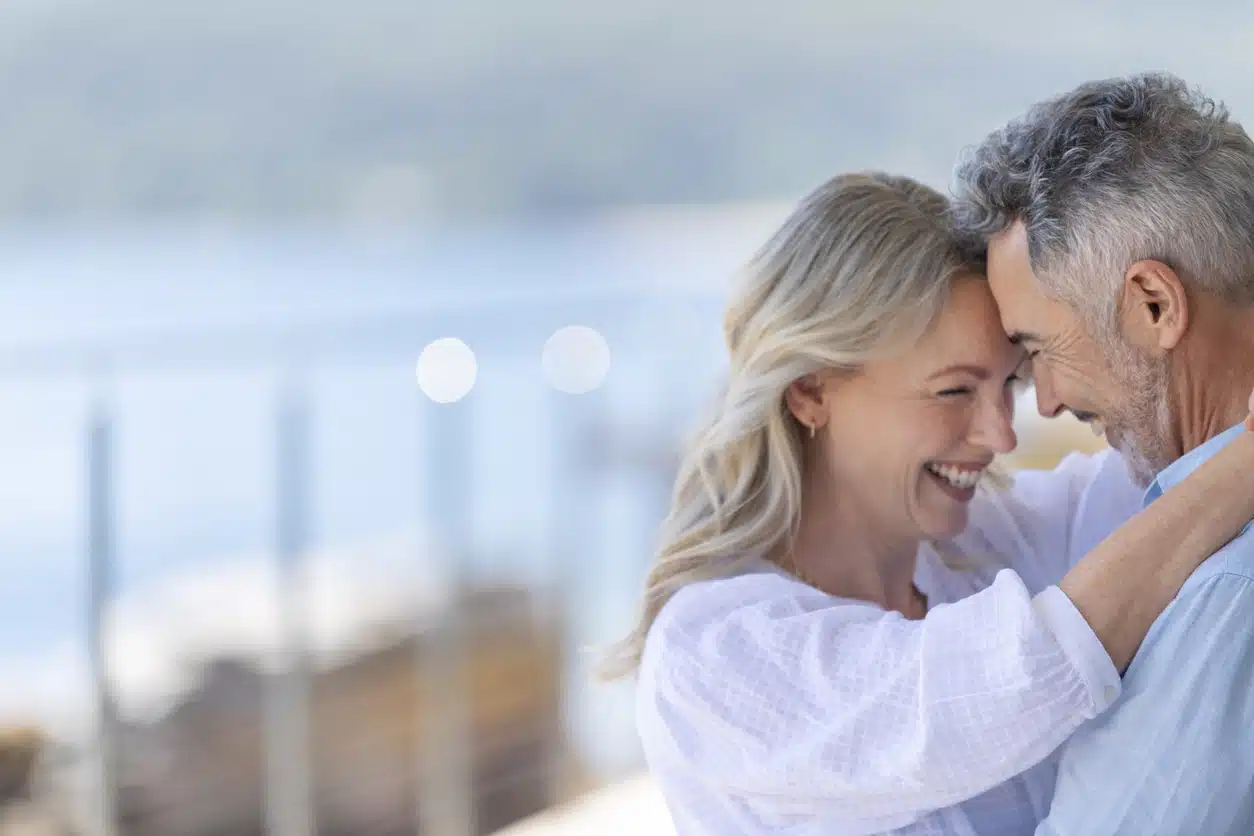 Mature couple dancing at a waterfront home or holiday villa. They are smiling and having fun. Probably on a romantic vacation. The woman has her arms around the man. The ocean is in the background