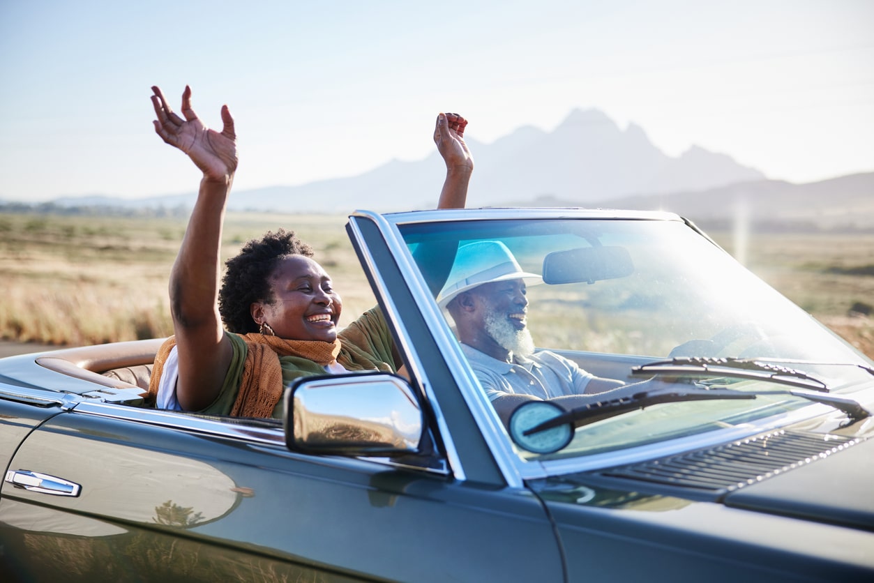 Mature African woman smiling with her hands raised in the air during a scenic road trip with her husband in their convertible