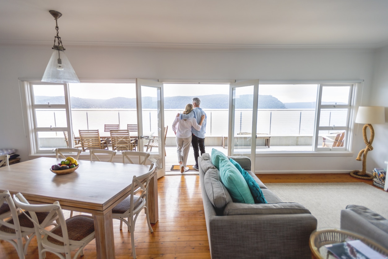 Mature couple looking at the view in their waterfront home. They look happy and contented. They are embracing. The ocean can be seen in the background.