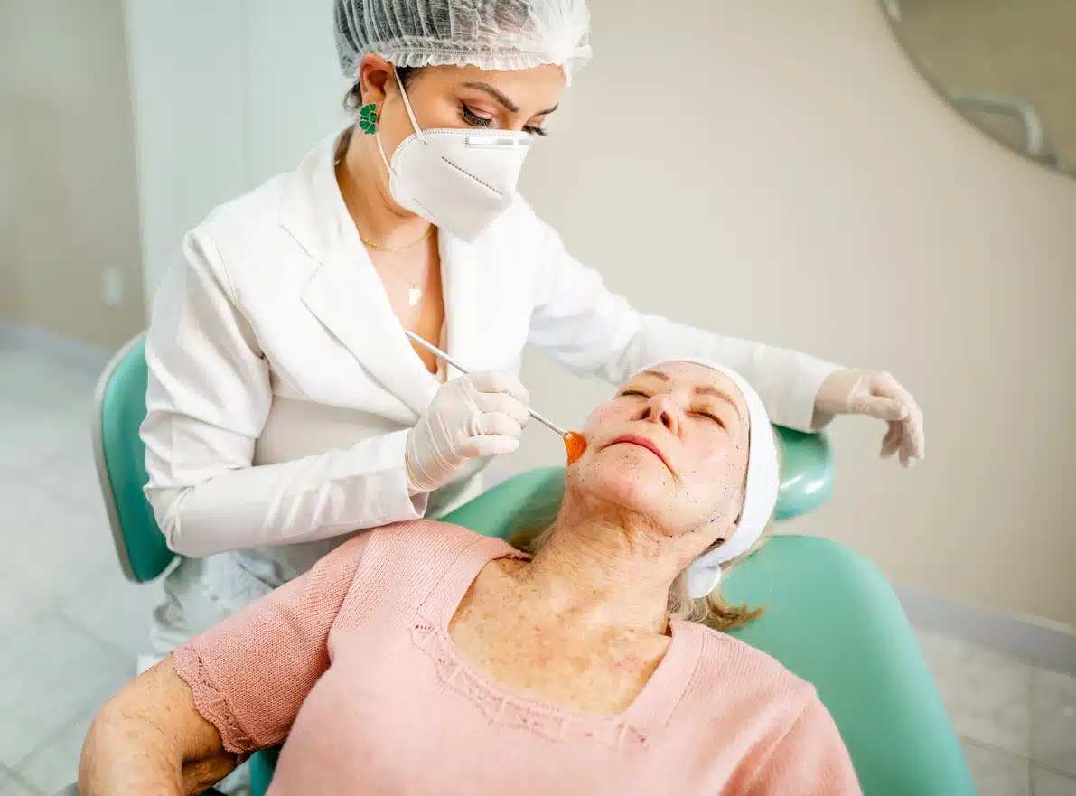 Beautician wearing protective workwear brushing numbing cream on a senior patient's face before a botox treatment