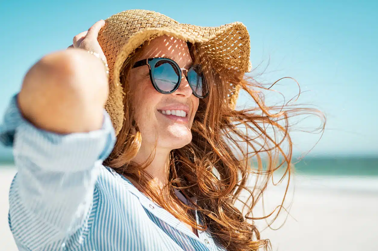 Side view of beautiful mature woman wearing sunglasses enjoying at beach. Young smiling woman on vacation looking away while enjoying sea breeze wearing straw hat. Closeup portrait of attractive girl relaxing at sea.