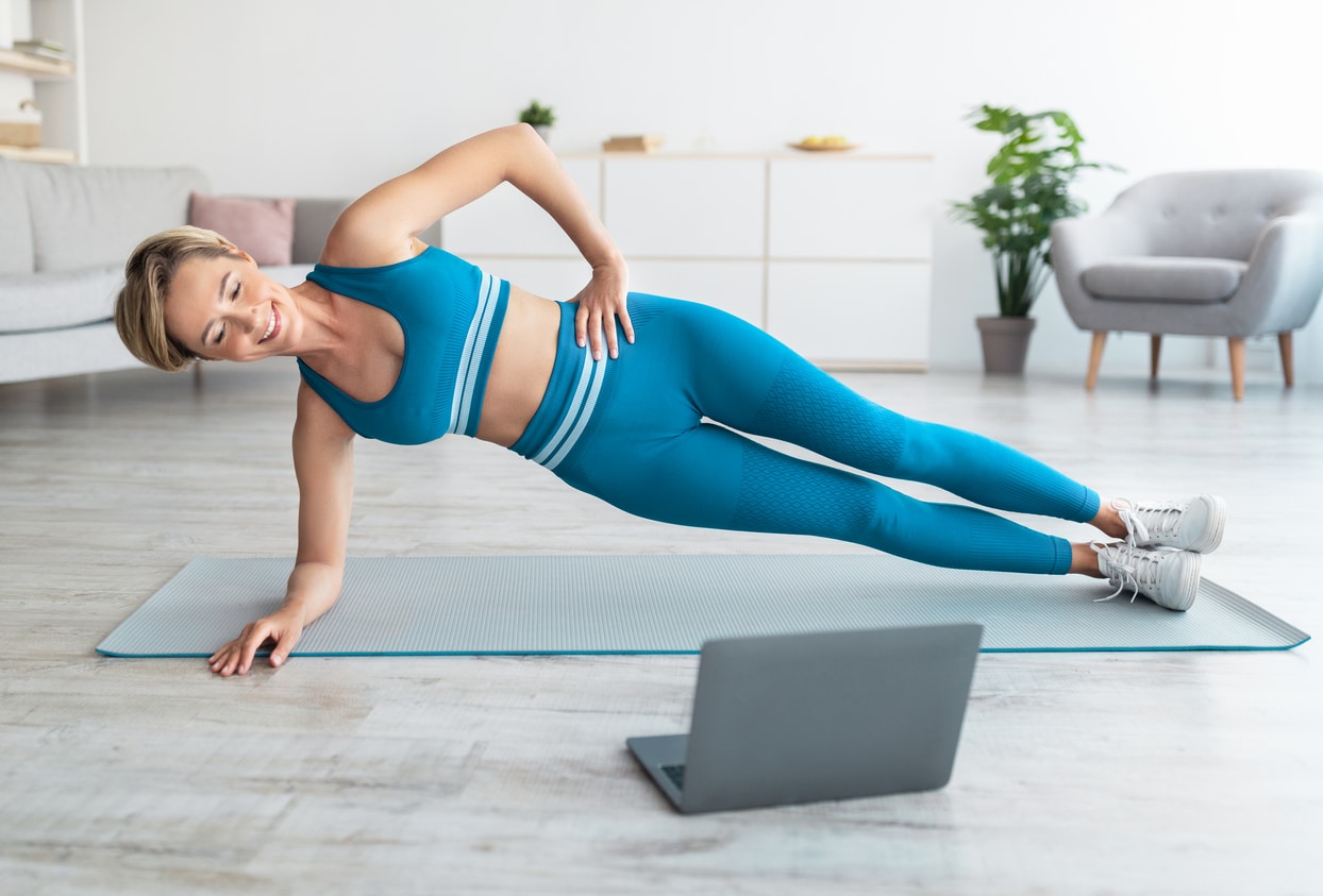Full Body Length Of Happy Fit Woman Exercising Standing In Side Plank Position On Floor Yoga Mat In Living Room, Looking At Computer Screen Watching Tutorial Or Virtual Workout With Trainer