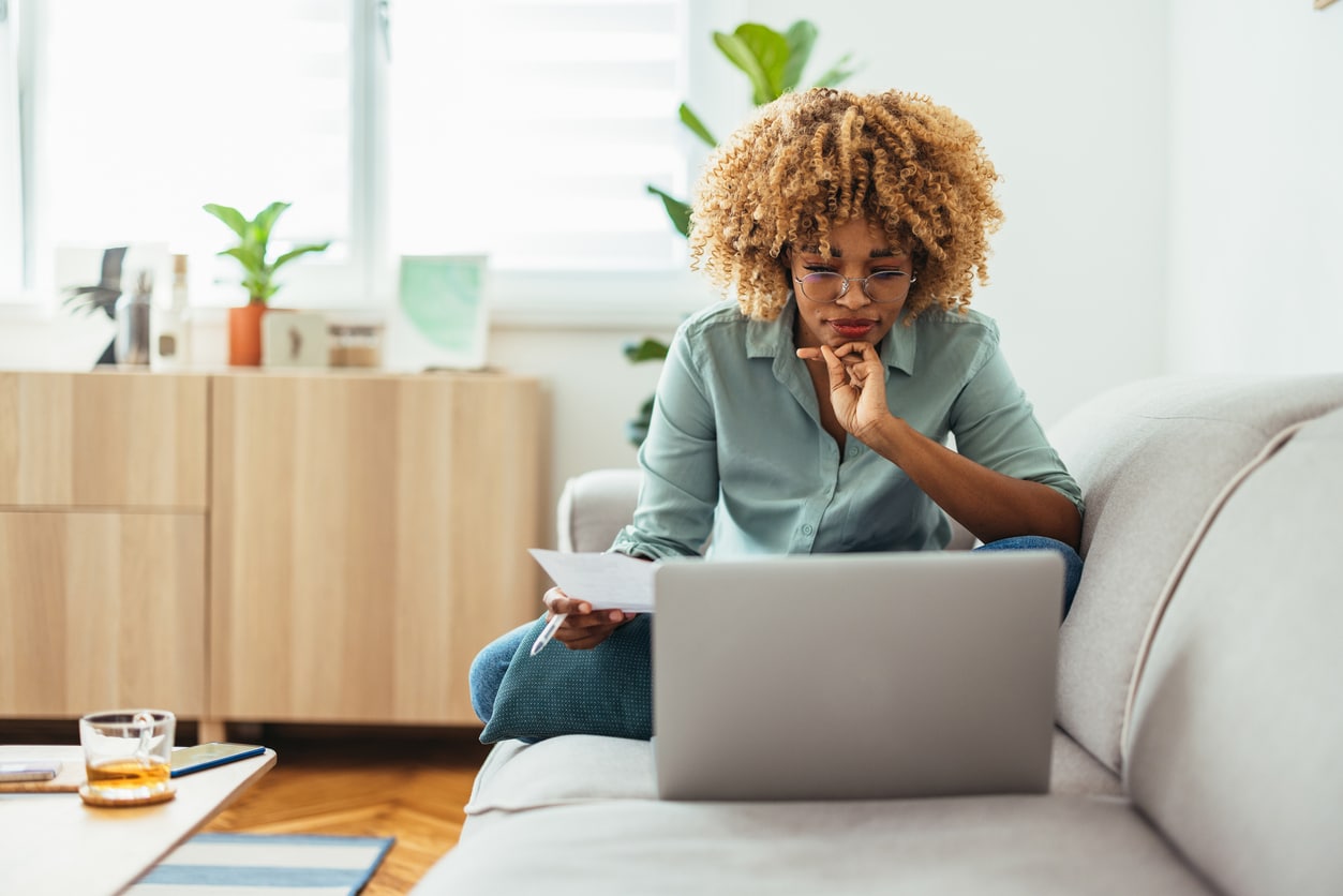 Serious Afro-American woman is sitting on a sofa and looking at a laptop while holding a paper in her hand. She is wearing glasses. She might be working from home or studying, or paying a utility bill. She is wearing a green shirt and jeans.