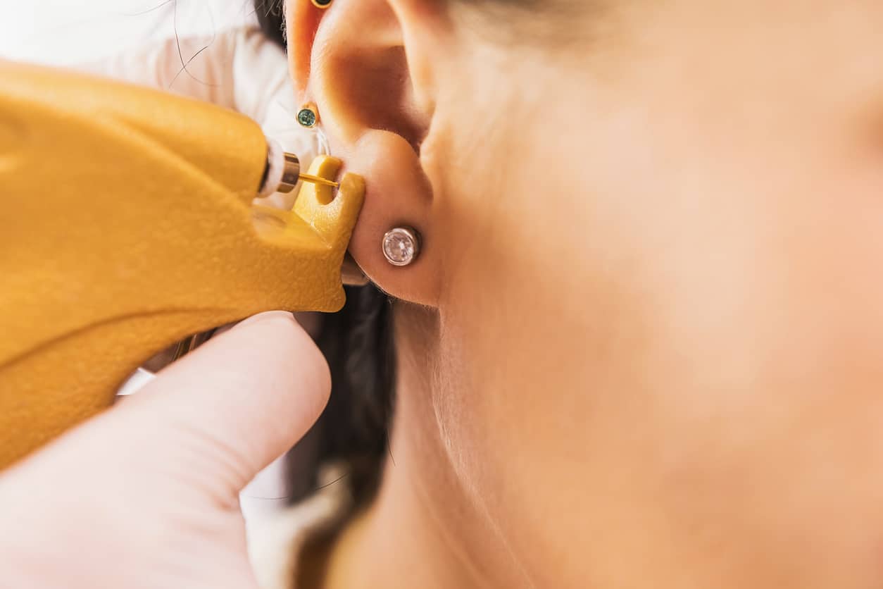 A doctor in sterile white medical gloves pierces the ears of a young girl in the medical office with a piercing gun.