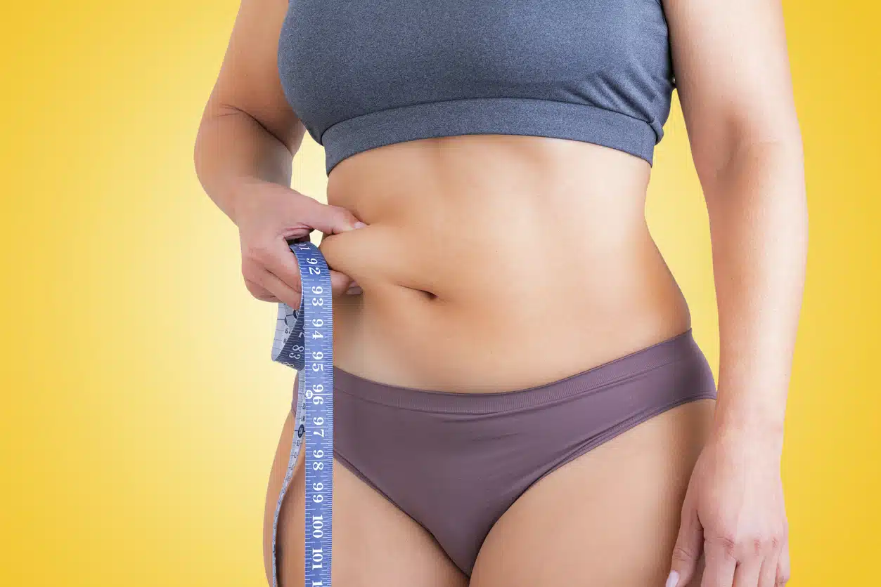 Healthcare and medicine: side view of unrecognizable woman squeezing her waist with fingers showing fat excess shot against yellow background.