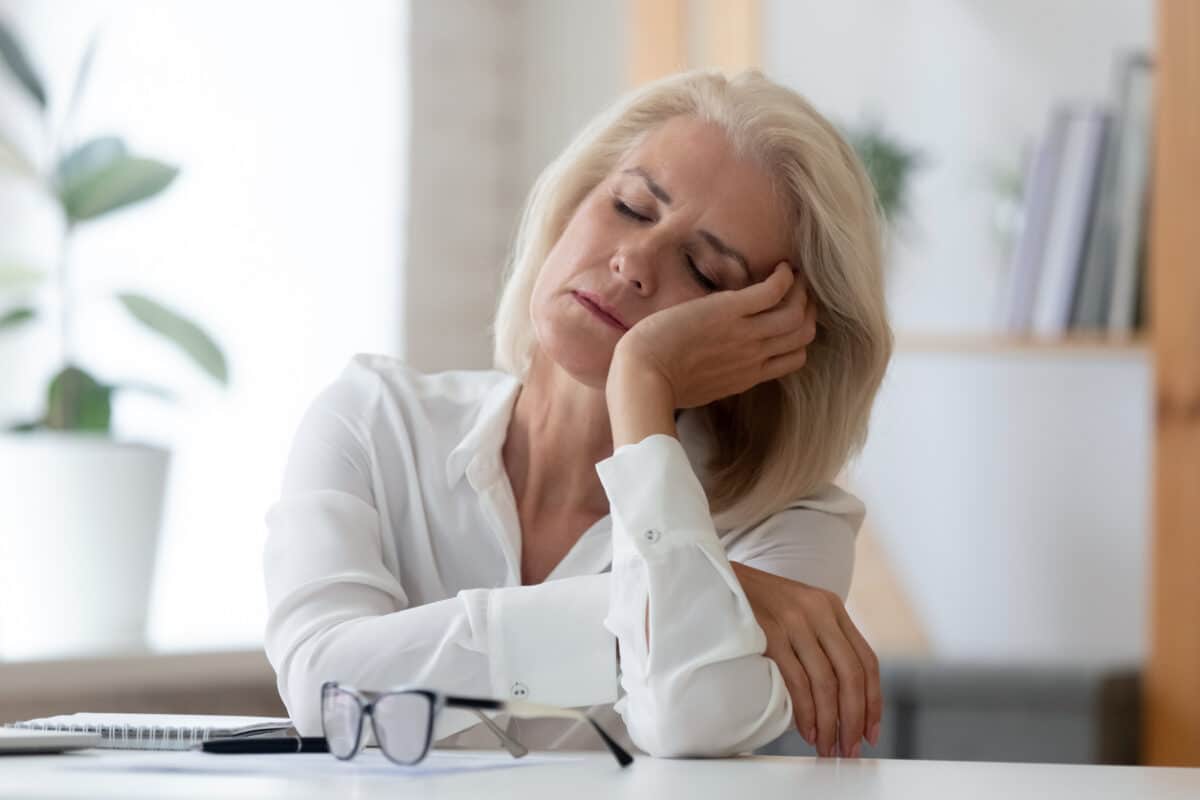 Exhausted aged woman worker sit at office desk fall asleep distracted from work, tired senior businesswoman feel fatigue sleeping at workplace taking break dreaming or visualizing
