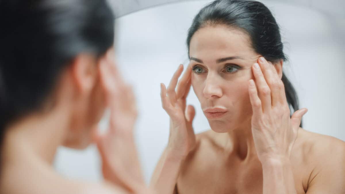 Caucasian Woman Looking in Mirror, Touching Beautiful Face, Soft Skin. Elderly Lady with Lush Black Hair. Female Beauty, Dignity, Grace. Sensual Wellness and Organic Skincare Products. Portrait Shot