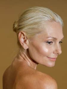 Studio portrait of graceful mature blonde woman with perfect glowing skin and natural makeup looking aside with a smile, posing naked isolated over beige background. Self acceptance, beauty concept