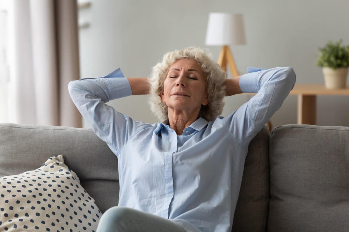 70s woman dreamer seated on couch relaxing meditating thinking positive thoughts putting hands behind head closed eyes breathing fresh air relish retired life free time at home, well-being concept