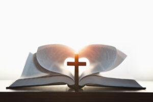 A cross symbolizing the death and resurrection of Jesus and a brightly lit heart-shaped Bible, the love of Jesus Christ in the background. Are We Loved? What Scripture Says