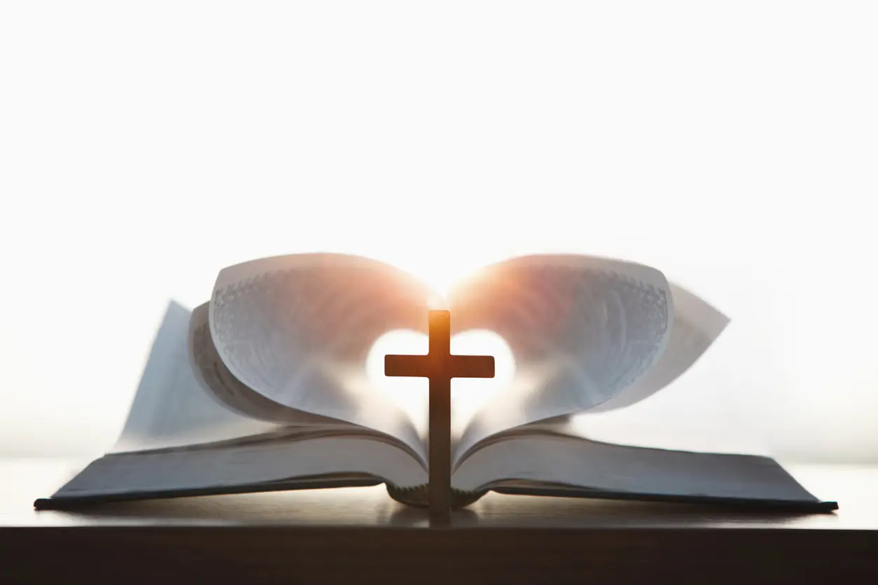 A cross symbolizing the death and resurrection of Jesus and a brightly lit heart-shaped Bible, the love of Jesus Christ in the background. Are We Loved? What Scripture Says
