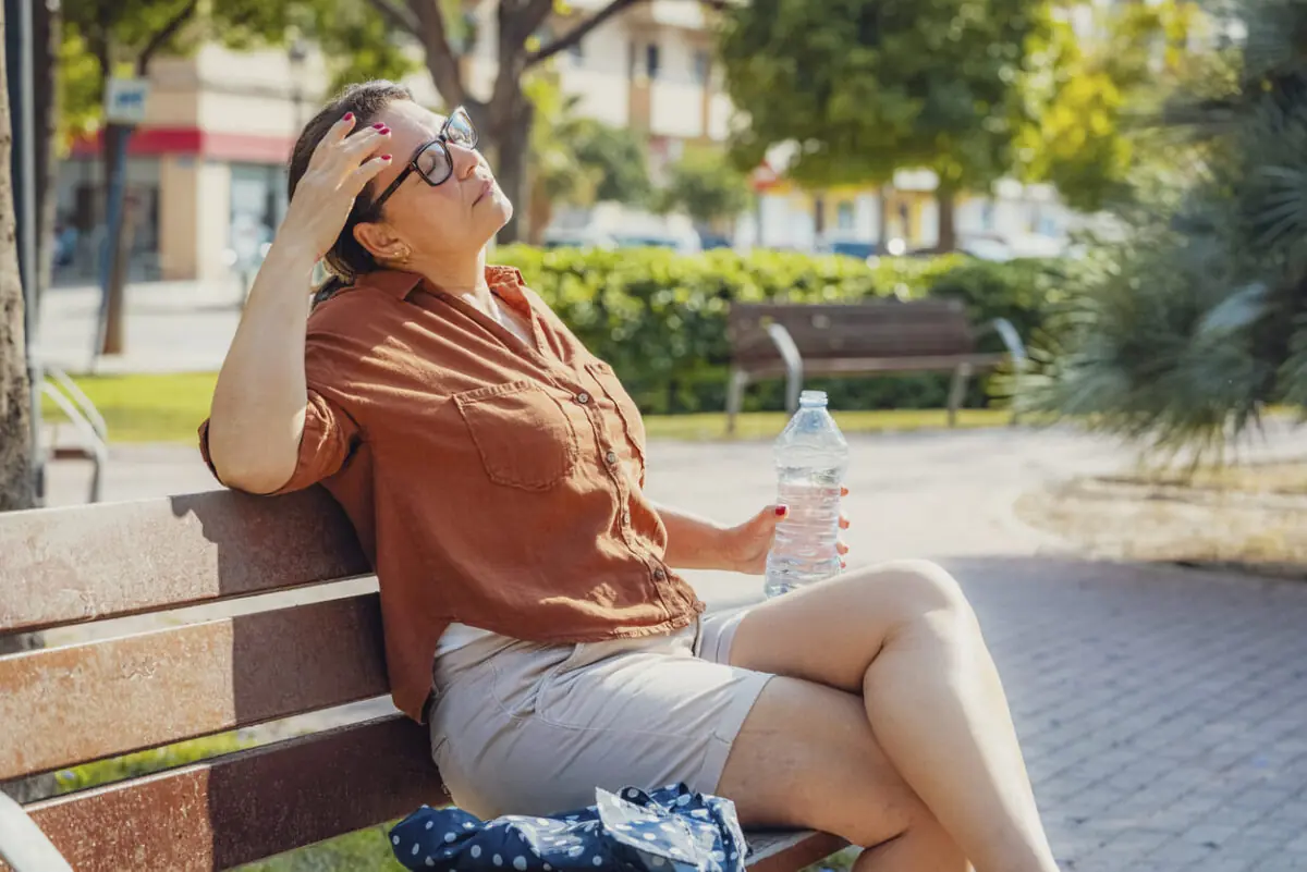 Woman resting on a park bench in bright sunlight, holding a bottle of water listening to the surroundings.